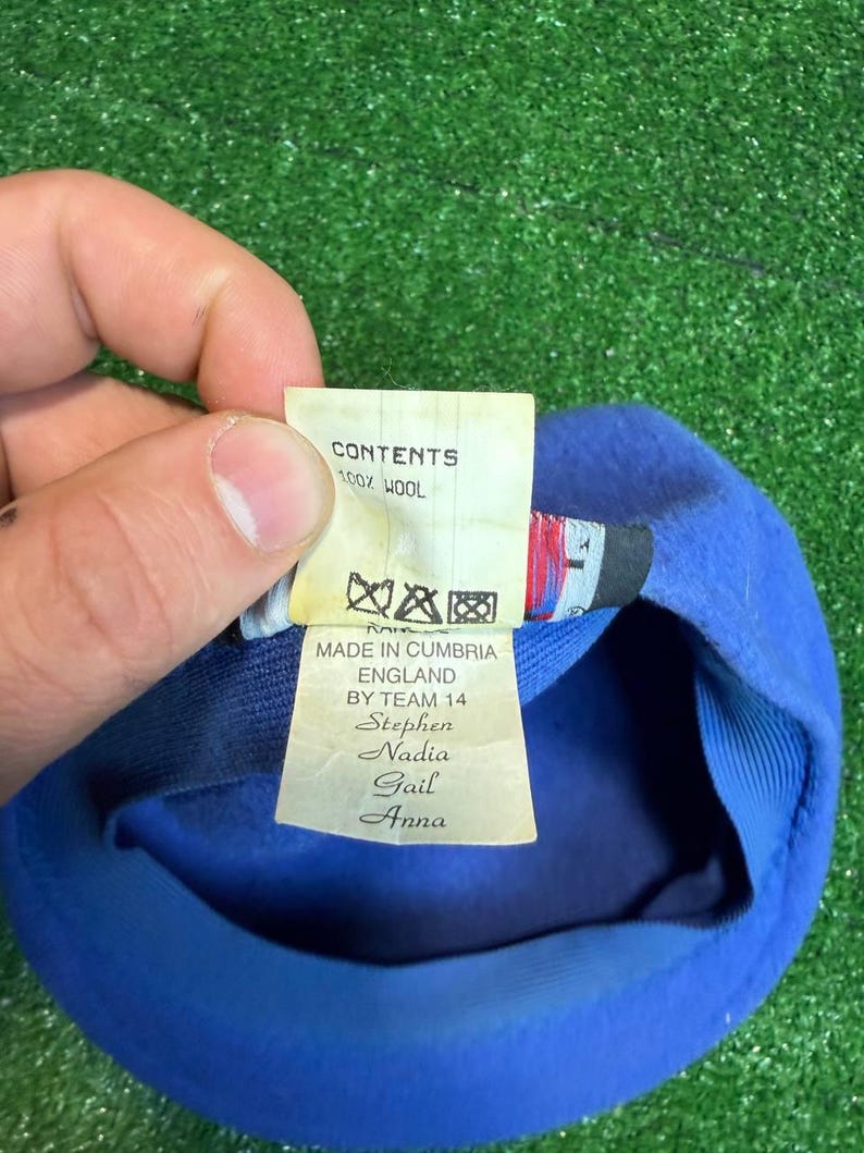K&ouml;nnte beinhalten: Blauer Wollbarett mit einem Etikett mit der Aufschrift "CONTENTS 100% WOOL" und "MADE IN CUMBRIA ENGLAND BY TEAM 14 Stephen Nadia Gail Anna". Der Barett liegt auf einer gr&uuml;nen Oberfl&auml;che.