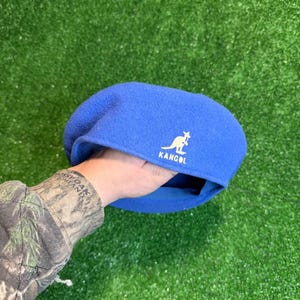 K&ouml;nnte beinhalten: Ein blauer Kangol-Barett mit wei&szlig;em K&auml;nguru-Logo und Markennamen. Der Barett wird vor einem gr&uuml;nen Hintergrund gehalten. Der Barett ist ein klassischer Stil aus weichem Material.