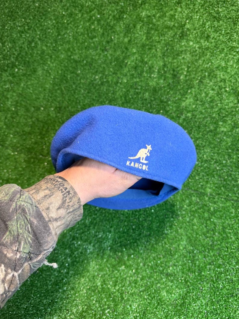 K&ouml;nnte beinhalten: Ein blauer Kangol-Barett mit wei&szlig;em K&auml;nguru-Logo und Markennamen. Der Barett wird vor einem gr&uuml;nen Hintergrund gehalten. Der Barett ist ein klassischer Stil aus weichem Material.