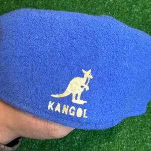 K&ouml;nnte beinhalten: Blauer Kangol-Barett mit wei&szlig;em K&auml;nguru-Logo und Markennamen. Der Hut ist aus einem weichen, filzartigen Material gefertigt. Der Barett ist ein klassischer Stil, der f&uuml;r verschiedene Anl&auml;sse geeignet ist.