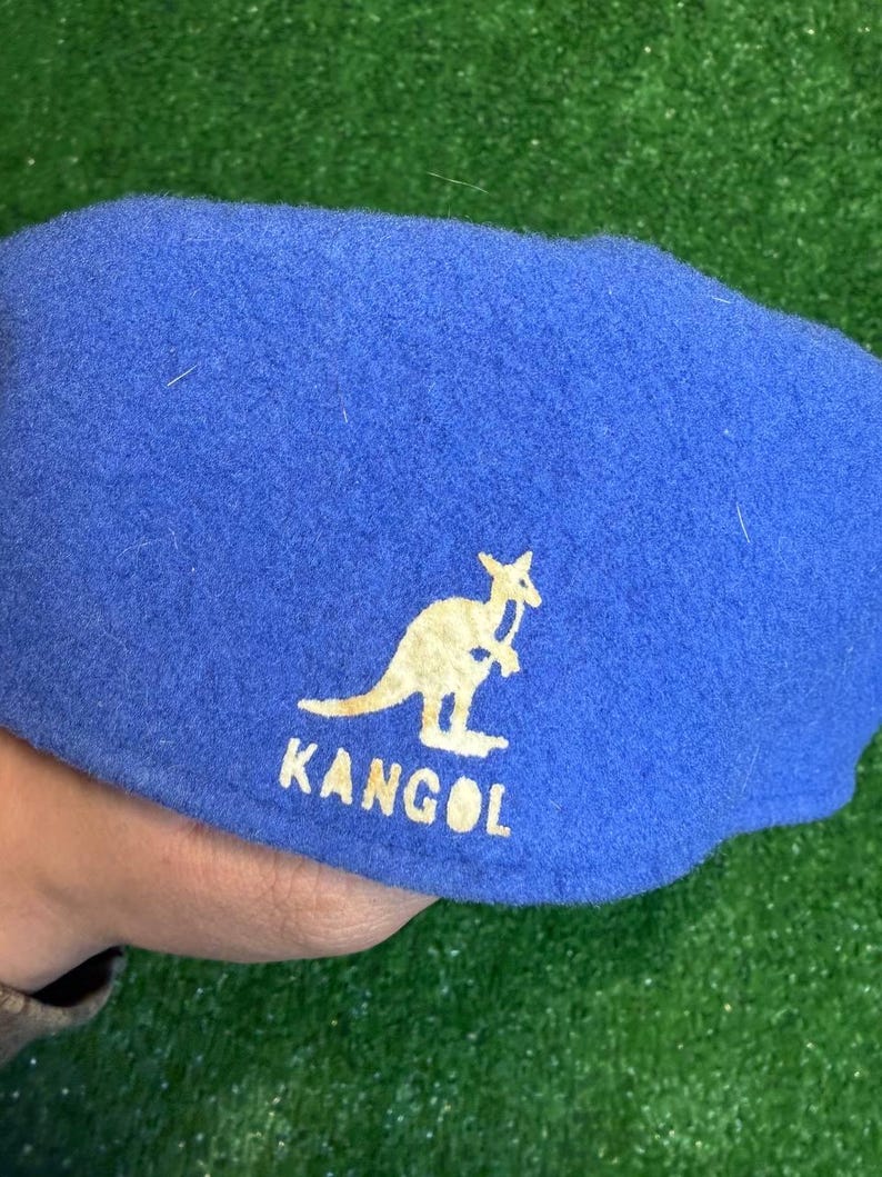 K&ouml;nnte beinhalten: Blauer Kangol-Barett mit wei&szlig;em K&auml;nguru-Logo und Markennamen. Der Hut ist aus einem weichen, filzartigen Material gefertigt. Der Barett ist ein klassischer Stil, der f&uuml;r verschiedene Anl&auml;sse geeignet ist.