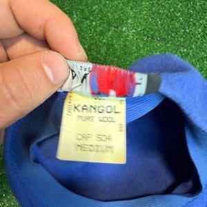 K&ouml;nnte beinhalten: Ein blauer Kangol-Hut mit einem Etikett mit der Aufschrift "KANGOL PURE WOOL CAP 504 MEDIUM". Der Hut ist aus weichem Material gefertigt und hat ein klassisches Design. Das Etikett ist wei&szlig; mit schwarzem Text und roten Akzenten.