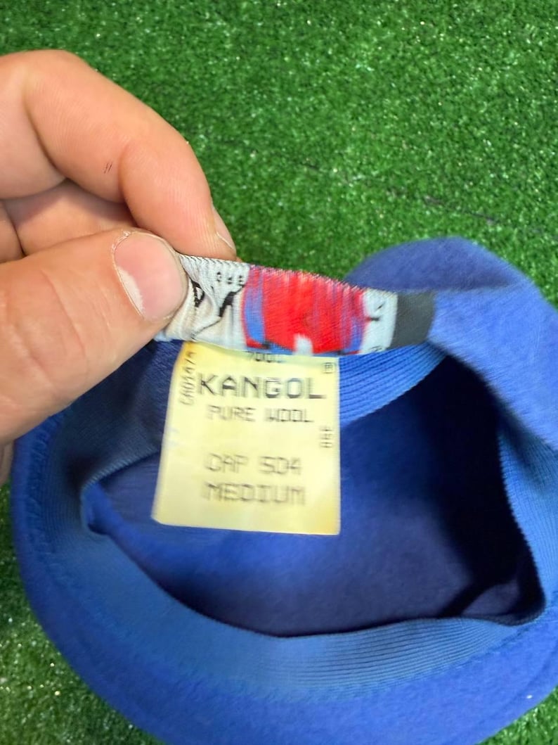 K&ouml;nnte beinhalten: Ein blauer Kangol-Hut mit einem Etikett mit der Aufschrift "KANGOL PURE WOOL CAP 504 MEDIUM". Der Hut ist aus weichem Material gefertigt und hat ein klassisches Design. Das Etikett ist wei&szlig; mit schwarzem Text und roten Akzenten.