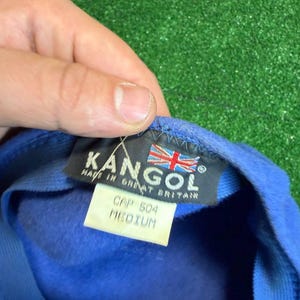 K&ouml;nnte beinhalten: Blaue Kangol-Kappe mit einem Etikett mit der Aufschrift "CAP 504 MEDIUM" und "MADE IN GREAT BRITAIN". Die Kappe hat das Kangol-Logo und eine britische Flagge. Die Kappe wird von einer Hand gehalten.