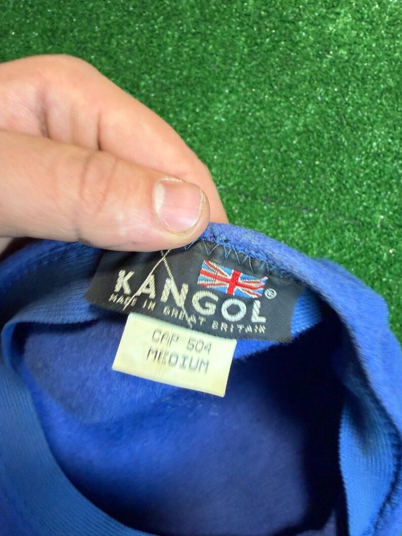 K&ouml;nnte beinhalten: Blaue Kangol-Kappe mit einem Etikett mit der Aufschrift "CAP 504 MEDIUM" und "MADE IN GREAT BRITAIN". Die Kappe hat das Kangol-Logo und eine britische Flagge. Die Kappe wird von einer Hand gehalten.