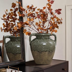 Könnte beinhalten: Eine große, grüne Keramikvase mit zwei Henkeln, gefüllt mit Zweigen aus orangefarbenen und roten Blättern. Die Vase hat ein zweifarbiges Design, mit einer dunkelgrünen Oberseite und einer hellbraunen Basis. Die Vase steht auf einer dunkelbraunen Oberfläche.