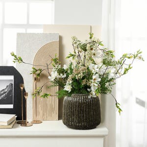 Könnte beinhalten: Dekorative Anordnung mit einer dunkelbraunen, strukturierten Vase, gefüllt mit weißen Blumen und Grün. Die Vase steht auf einer weißen Oberfläche, mit einem gerahmten Schwarz-Weiß-Bergfoto, goldenen Kerzenhaltern und einem strukturierten Wandkunstwerk im Hintergrund.