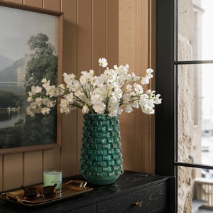 Könnte beinhalten: Eine türkisfarbene Keramikvase mit gewebter Textur hält einen Strauß weißer Blumen. Die Vase steht auf einer schwarzen Kommode, neben einem gerahmten Landschaftsbild und einem goldenen Tablett mit Accessoires.