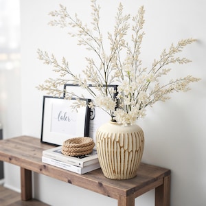 Könnte beinhalten: Eine cremefarbene Keramikvase mit vertikalen braunen Linien hält einen Strauß weißer Blumen und Zweige. Die Vase steht auf einer Holzbank mit Büchern, Untersetzern und gerahmter Kunst. Die gerahmte Kunst trägt das Wort "later".
