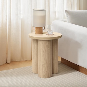 Könnte beinhalten: Ein Beistelltisch aus hellem Holz mit runder Tischplatte und drei zylindrischen Beinen. Auf dem Tisch stehen eine Lampe mit beigem Schirm und gesprenkeltem Sockel, ein klares Glas und ein Untersetzer. Der Tisch steht neben einem weißen Sofa.