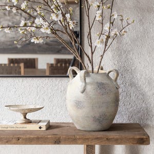 Könnte beinhalten: Eine große, cremefarbene Keramikvase mit zwei Henkeln, gefüllt mit Zweigen mit weißen Blüten. Die Vase steht auf einem rustikalen Holzregal, mit einer kleinen dekorativen Schale und einem Buch mit dem Titel "Vintage Table Decoration".