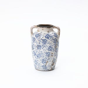 Könnte beinhalten: Eine weiße Keramikvase mit blauen Blumenmustern und zwei Henkeln. Die Vase hat ein abgenutztes, gealtertes Aussehen mit einem dunkelbraunen Rand und subtilen Unvollkommenheiten, was eine Vintage-Ästhetik suggeriert.