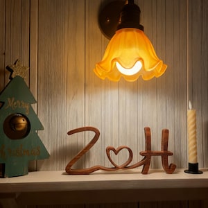 Puede incluir: Una l&aacute;mpara de pared de tono c&aacute;lido con una pantalla de vidrio &aacute;mbar con volantes ilumina una estanter&iacute;a. Un recorte de madera que deletrea "ZoH" con un detalle de coraz&oacute;n se encuentra en la estanter&iacute;a, junto con una decoraci&oacute;n de &aacute;rbol de Navidad y una vela.