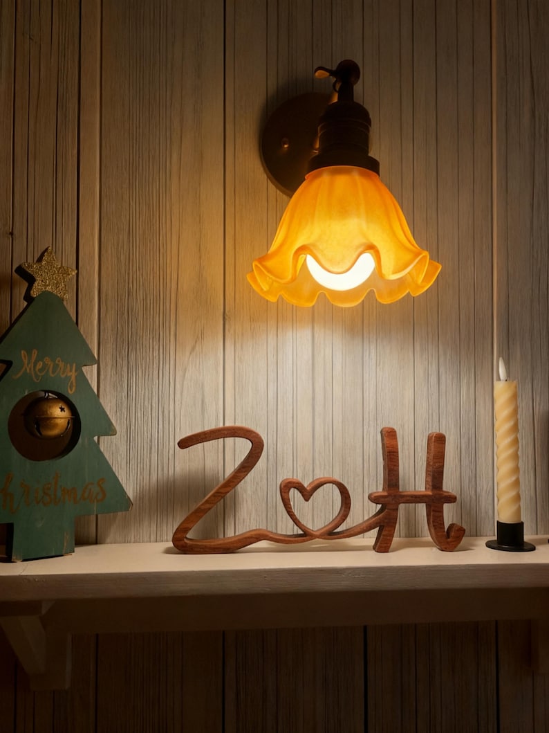 Puede incluir: Una l&aacute;mpara de pared de tono c&aacute;lido con una pantalla de vidrio &aacute;mbar con volantes ilumina una estanter&iacute;a. Un recorte de madera que deletrea "ZoH" con un detalle de coraz&oacute;n se encuentra en la estanter&iacute;a, junto con una decoraci&oacute;n de &aacute;rbol de Navidad y una vela.