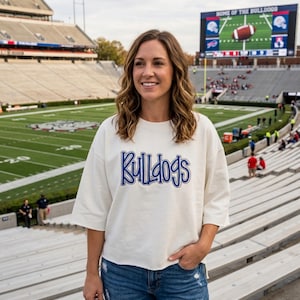 May include: An off-white, short-sleeved sweatshirt featuring the word Bulldogs in blue, outlined lettering across the chest, paired with blue denim jeans. The garment has a relaxed fit with a straight hemline and a crew neckline.