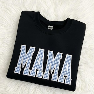 Könnte beinhalten: Schwarzes Sweatshirt mit dem Wort "MAMA" in großen, weißen Blockbuchstaben. Die Buchstaben sind mit einem blau-weißen Vichy-Muster gefüllt. Das Sweatshirt wird auf einer weißen, flauschigen Oberfläche präsentiert.