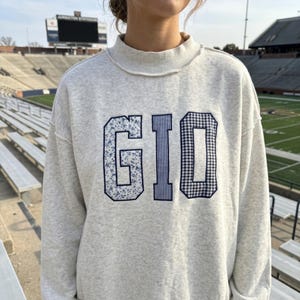May include: A light gray heathered sweatshirt with a mock neck and raw-edge hem. The front features the letters GIO in large, dark blue-outlined applique patches, with the G in a floral print, the I in vertical stripes, and the O in gingham.