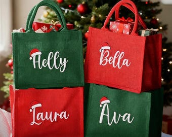 Könnte beinhalten: Vier gestapelte Weihnachts-Tragetaschen in Rot und Grün. Jede Tasche hat einen Namen in weißer Schrift mit einem Weihnachtsmannhut. Die Taschen sind mit Geschenken gefüllt, und im Hintergrund ist ein Weihnachtsbaum zu sehen.