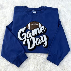 Peut inclure: Un sweat-shirt bleu marine avec l'inscription "Game Day" en blanc, soulignée de noir, et un motif de ballon de football marron et blanc. Le t-shirt à manches longues est en matière douce.
