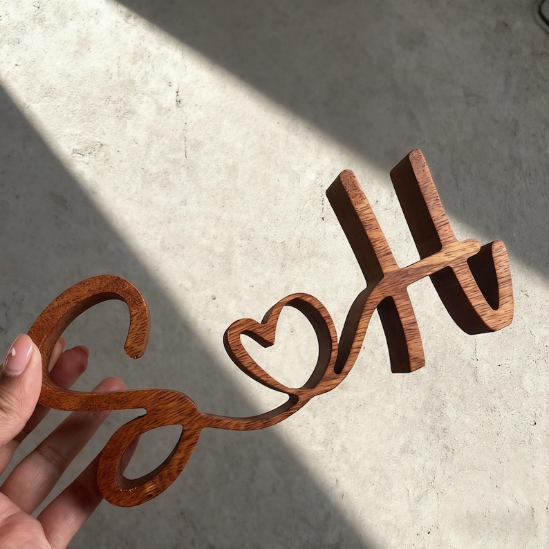 Puede incluir: Letrero de madera 'So & H' en fuente cursiva, con un coraz&oacute;n reemplazando la 'o'. Las letras son de color marr&oacute;n c&aacute;lido, y el letrero se sostiene contra un fondo neutro. El letrero es probablemente un art&iacute;culo decorativo.