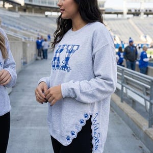 Könnte beinhalten: Ein hellgraues Langarm-Sweatshirt mit einem blau-weiß karierten KY-Logo auf der Brust und dem Wort Wildcats in blauer Schreibschrift. Der Saum hat einen geschwungenen Seitenschlitz mit blauen Pfotenabdrücken und dem vertikal gedruckten Wort Wildcats.