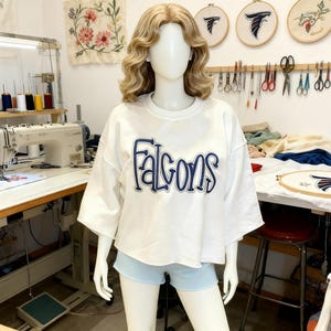 May include: A white, short-sleeved sweatshirt with the word "Falcons" in blue script across the front. The sweatshirt is displayed on a mannequin. The background includes sewing equipment and embroidered wall hangings.