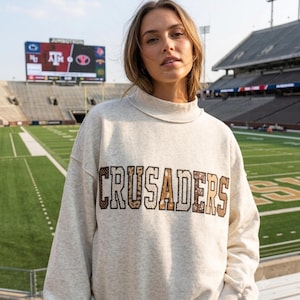 Op de afbeelding: Een langarmig sweatshirt in een gemêleerde haverkleur met een opstaande kraag. Op de voorzijde staat het woord CRUSADERS in grote blokletters, elk gevuld met een variëteit aan bruine en beige dierenprints en stippenpatronen.