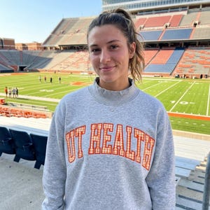 Op de afbeelding: Een grijs gemêleerd sweatshirt met lange mouwen en een opstaande kraag. Op de voorzijde staat de tekst UT HEALTH in oranje en wit geblokte letters met een dunne oranje omlijning, gecentreerd op de borst.