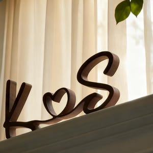 Puede incluir: Iniciales de madera "K & S" en fuente cursiva, con un coraz&oacute;n reemplazando la & comercial. Las letras son de color marr&oacute;n c&aacute;lido, sobre un fondo neutro de ventana y cortinas. Un art&iacute;culo decorativo para la decoraci&oacute;n del hogar.