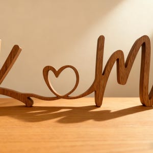 Puede incluir: Pieza decorativa de madera con las letras "K" y "M" unidas por un coraz&oacute;n. Las letras y el coraz&oacute;n est&aacute;n hechos de madera de tonos c&aacute;lidos, proyectando sombras sobre la superficie. Un dise&ntilde;o sencillo y elegante.