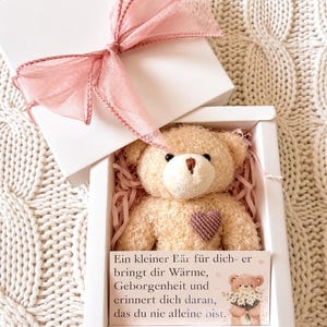 Könnte beinhalten: Ein beiger Teddybär mit einem kleinen dunklen Herz auf der Brust, in einer weißen Geschenkbox mit rosa Papierfüllung. Die Box ist mit einer rosa Schleife verziert und enthält eine Karte mit dem Text: Ein kleiner Bär für dich- er bringt dir Wärme.