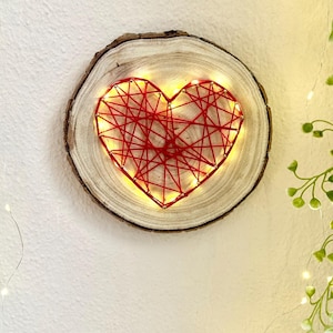 May include: A wooden slice with a red string heart design illuminated by warm white lights. The heart is centered on the wood, and the lights create a soft glow. The wood has a natural bark edge.