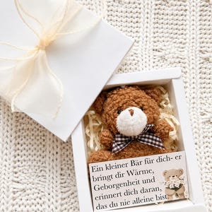 Könnte beinhalten: Ein brauner Teddybär mit weißer Schnauze und karierter Schleife, präsentiert in einer weißen Geschenkbox mit cremefarbenem Band. Die Box enthält zerknülltes Papier als Polsterung. Eine Karte mit deutschem Text ist sichtbar.