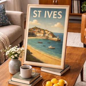 May include: A framed art print of St Ives, featuring a coastal town scene with a beach, boats, and a lighthouse. The print has a light wood frame and is displayed on a wooden coffee table with books, a mug, and a bowl of lemons.