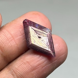 May include: A small, diamond-shaped crystal with a deep purple hue and iridescent gold and white internal striations. The crystal is held between fingers, showcasing its geometric form and natural beauty. The crystal's facets catch the light, creating a shimmering effect.