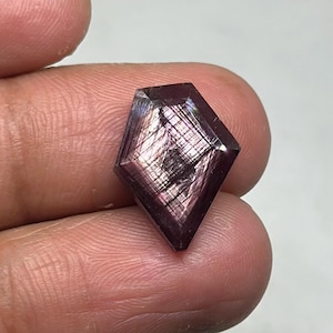 May include: A faceted, dark purple gemstone with a kite-like shape. The gem has a reflective, iridescent quality with internal lines and patterns. The stone is held between fingers, showcasing its size and detail.