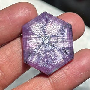 May include: A hexagonal, translucent purple gemstone featuring a distinct six-rayed star pattern across its surface. The stone displays a fibrous, textured appearance with varying shades of violet and light pink, showcasing a natural crystalline structure.