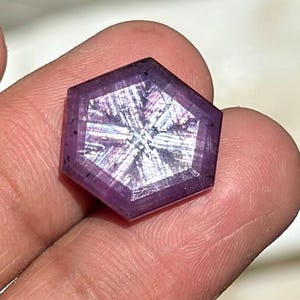 Puede incluir: Una gema hexagonal facetada de color púrpura intenso. La gema presenta un patrón en forma de estrella en su interior, que refleja la luz. La piedra se sostiene entre los dedos, mostrando su tamaño y detalle. Esta gema es adecuada para la fabricación de joyas.