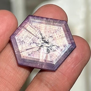 May include: A faceted, hexagonal gemstone with a purple hue. The gem has a complex internal pattern with a central dark area and radiating lines. The edges are a darker shade of purple, and the gem is held in a hand.