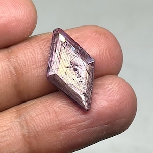 May include: A diamond-shaped gemstone with a mix of purple and gold hues. The gem has a polished surface and internal inclusions, creating a unique pattern. The gemstone is held in a hand.