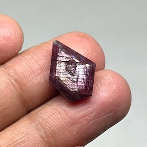 May include: A dark purple crystal with a geometric shape and a metallic sheen. The crystal is faceted, reflecting light and creating a shimmering effect. The crystal is held in a hand. The overall aesthetic is one of natural beauty and geological interest.