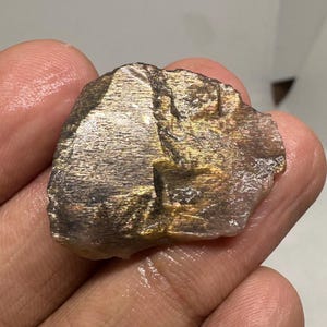 May include: A rough-cut, natural sunstone gemstone. The stone displays a mix of colors, including gold, brown, and hints of clear. The surface has a textured appearance, with visible crystalline structures. The stone is held in a hand.