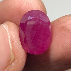 May include: A vibrant, oval-shaped, faceted gemstone with a deep magenta-pink hue. The stone features a polished surface with multiple geometric facets that reflect light, showcasing a translucent quality and a rich, saturated color throughout.