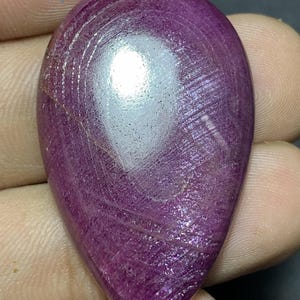 May include: A pear-shaped cabochon gemstone with a deep magenta and purple hue. The surface displays a smooth, polished finish with visible internal striations and a bright white light reflection centered on the upper portion of the stone.