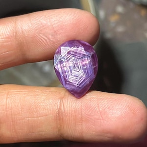 May include: A faceted, pear-shaped gemstone with a deep purple hue. The gem's interior features a complex, geometric pattern of light and dark purple lines, creating a starburst effect. The stone is held between two fingers.
