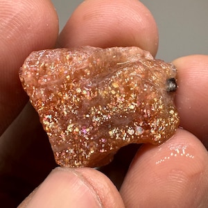 May include: A rough-cut sunstone gemstone with a sparkling, iridescent appearance. The stone has a warm, orange-brown base color with flecks of gold, pink, and other colors. The sunstone is held between fingers.