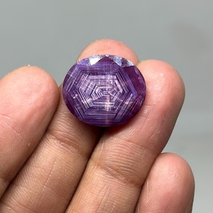 Puede incluir: Una gema púrpura redonda y facetada con un patrón de estrella. La gema se sostiene en una mano, mostrando su intrincado diseño y color. Las facetas de la gema capturan la luz, creando un efecto brillante.