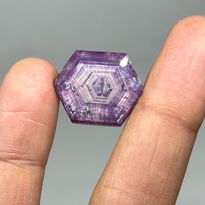 May include: A hexagonal, purple gemstone with a complex internal structure. The gem is held between two fingers, showcasing its intricate facets and color variations. The stone's surface reflects light, highlighting its crystalline patterns.