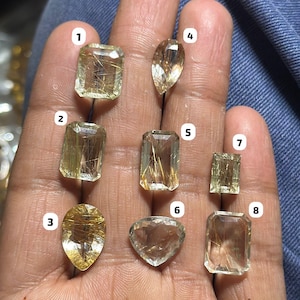 Puede incluir: Una colección de ocho gemas facetadas de varias formas, incluyendo rectangulares, ovaladas y en forma de pera. Las gemas son de color amarillo pálido con inclusiones doradas, sostenidas en una mano sobre un fondo de mezclilla azul. Las gemas están numeradas del 1 al 8.
