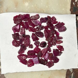 Puede incluir: Un conjunto de gemas de rubí en bruto, de color rojo intenso. Las piedras sin tallar, de formas y tamaños variados, se presentan sobre una superficie blanca, mostrando su belleza natural.